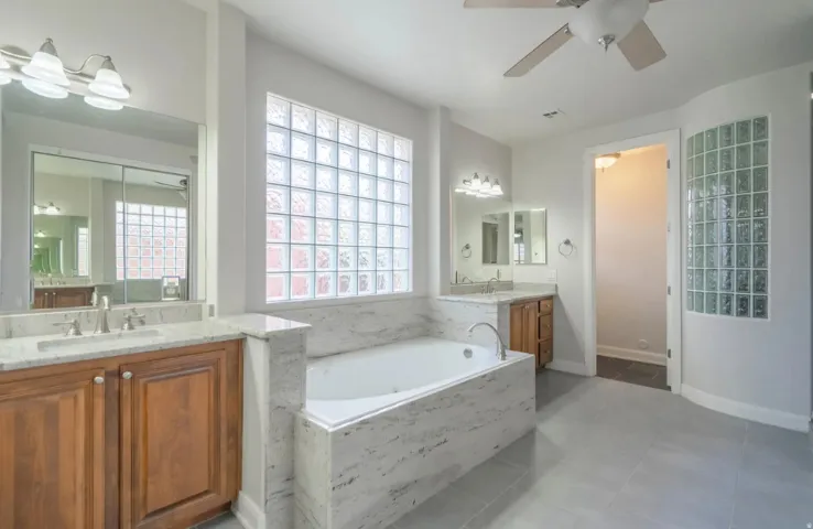 Light-filled bathroom featuring a soaking tub with stone surround, dual vanities with stone countertops, glass block window, and tile flooring