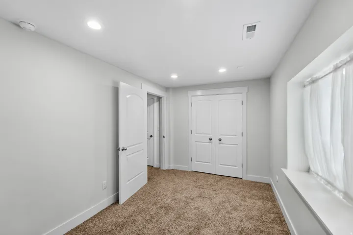 Comfortable basement bedroom bathed in soft light from recessed lighting and a charming window with sheer coverings. The peaceful neutral walls create a relaxing atmosphere, while plush soft carpeting enhances the cozy, inviting feel of this well-designed