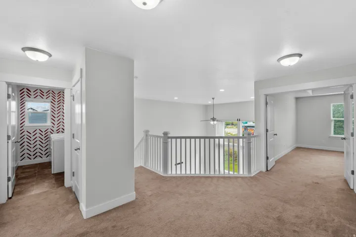 Attractive second-floor hallway connecting the bedrooms to the loft, featuring comfortable carpeting and a stylish white wood railing. The adjacent room's window and the laundry area's window flood the space with natural light, while a decorative patterne