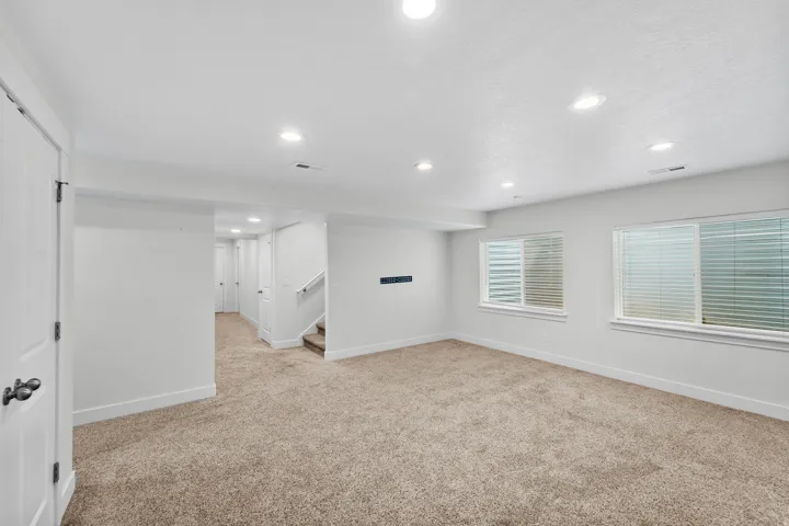 Spacious basement great room flooded with natural light from two windows and enhanced by recessed lighting for an airy, welcoming feel. Soft carpeted flooring creates a comfortable environment, while a stylish staircase with white railings seamlessly conn