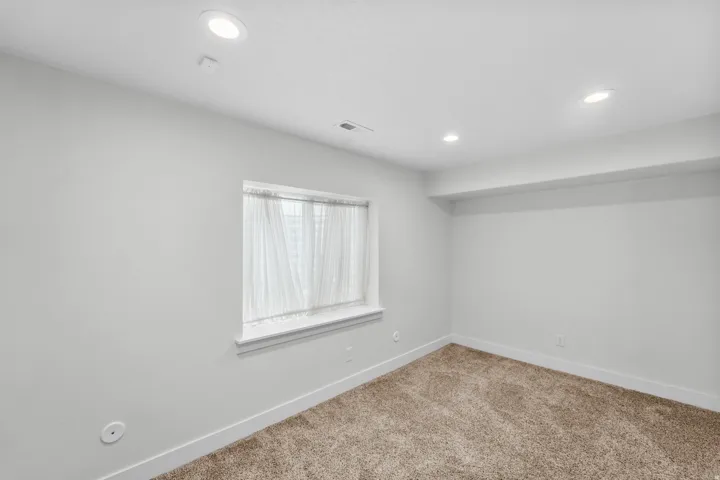 Comfortable basement bedroom bathed in soft light from recessed lighting and a charming window with sheer coverings. The peaceful neutral walls create a relaxing atmosphere, while plush soft carpeting enhances the cozy, inviting feel of this well-designed