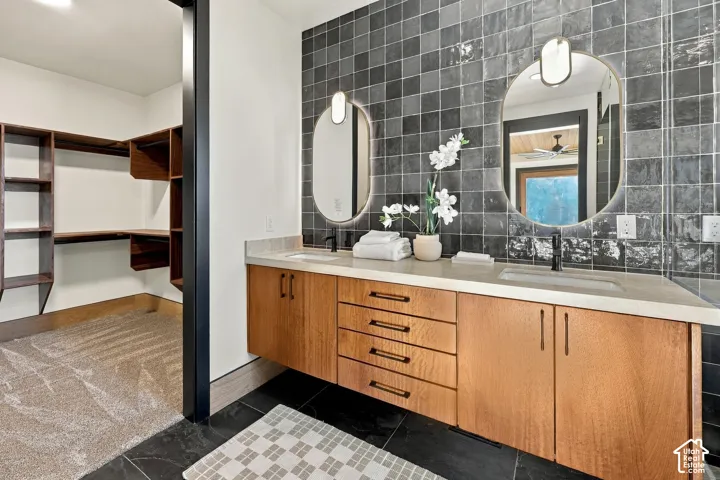 Master Ensuite with floating double vanity featuring wood-finish cabinetry, a neutral countertop with dual integrated sinks, matte black hardware, and two oval wall-mounted mirrors set against a dark square-tiled feature wall