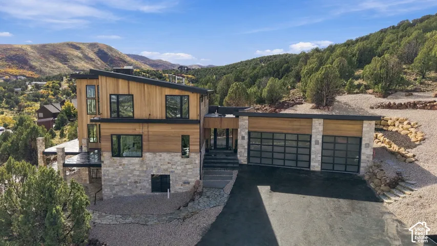 Contemporary multi-level exterior featuring wood siding, natural stone accents, shed-style roofing, and two glass-paneled garage doors, situated against a mountain backdrop