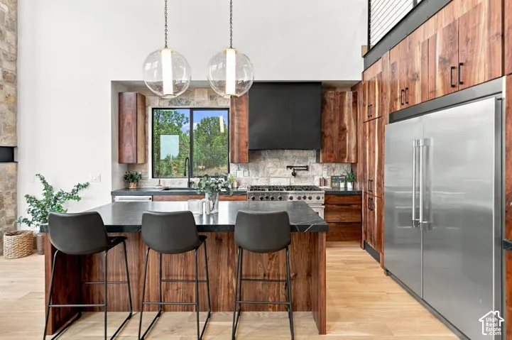 Kitchen featuring natural wood-finish cabinetry, stone backsplash, dark stone-top island, stainless steel refrigerator, and glass globe pendant lighting