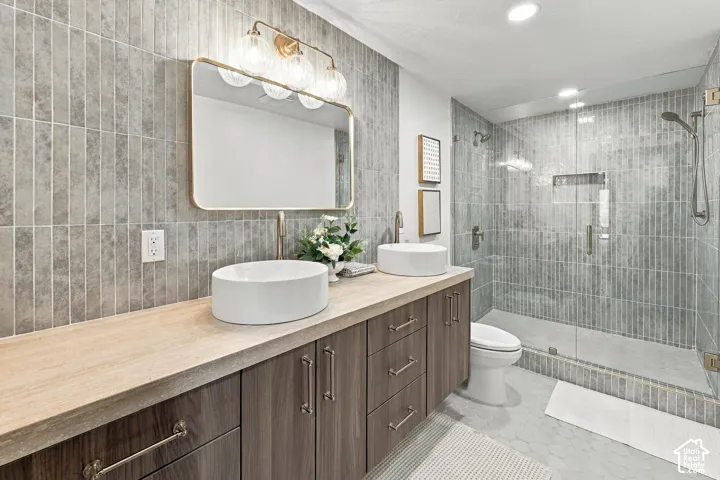 Dual-sink vanity with wood-finish cabinetry, countertop, circular vessel sinks, and brass-tone fixtures, complemented by vertical tile backsplash and a walk-in glass-enclosed shower with tiled surround