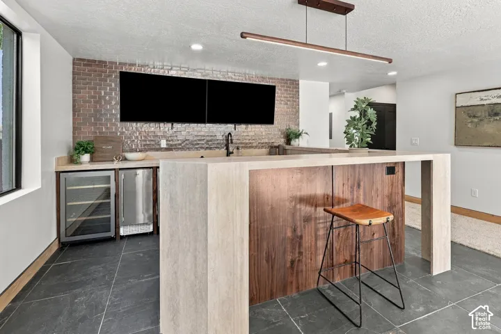 Custom wet bar featuring a brick tile backsplash, stone-finish waterfall countertop, built-in stainless steel beverage refrigerator, dark wood cabinetry, and linear pendant lighting