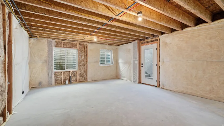 Unfinished basement space with exposed wood joists and subfloor, two windows with blinds, a glass-paneled door, and exposed insulation