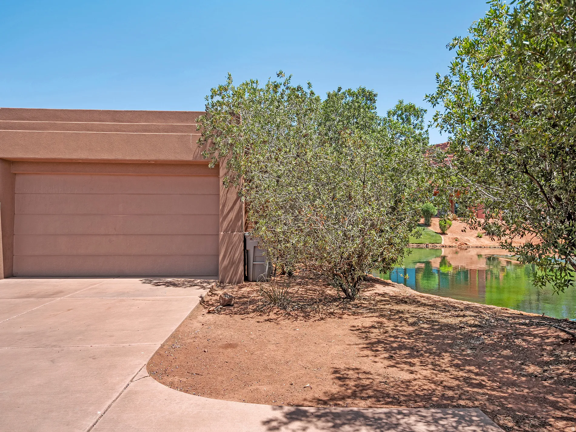 Stucco exterior with a garage door, featuring a concrete driveway, and natural landscaping with mature trees, bordering a tranquil body of water