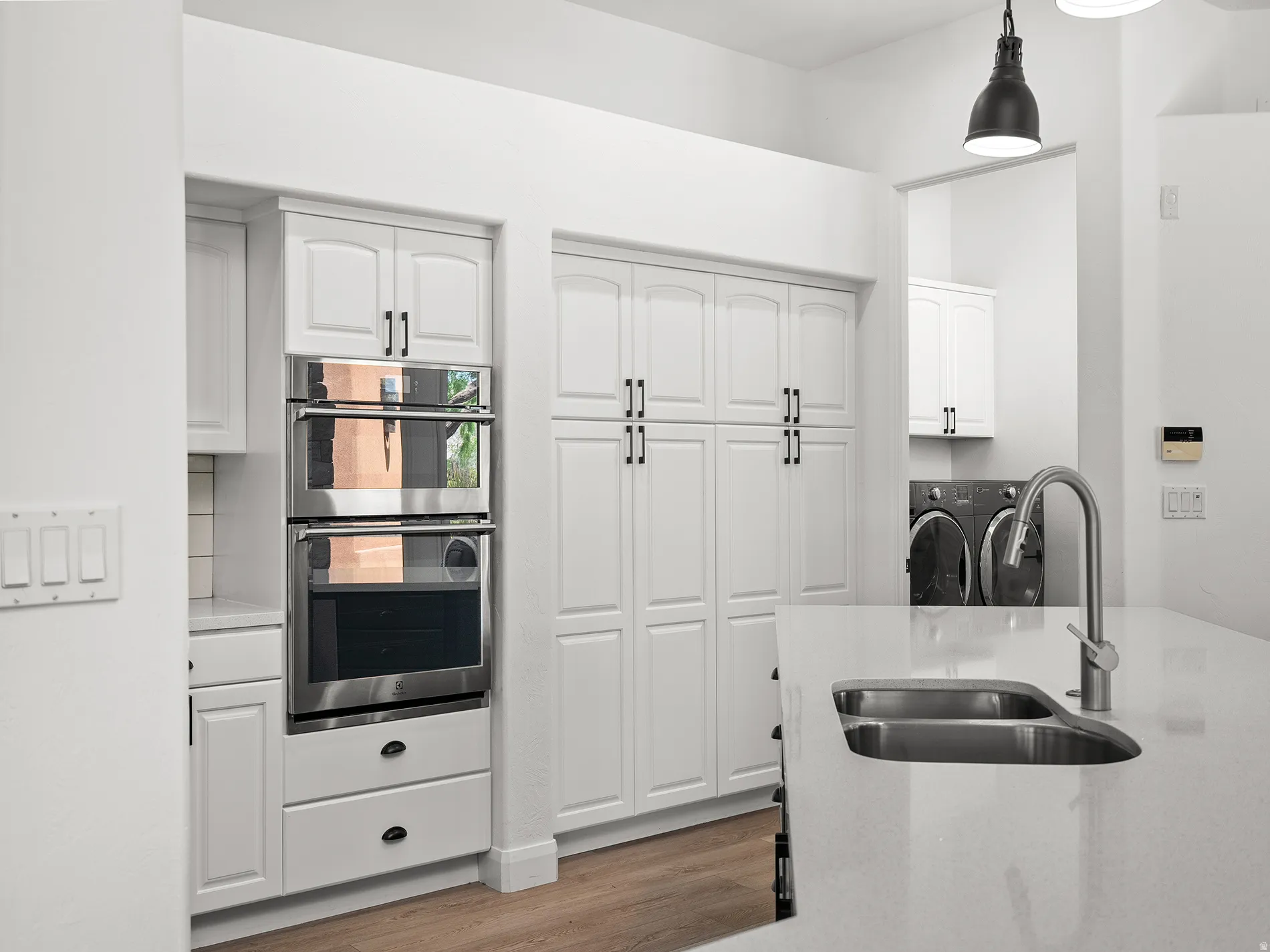 White cabinetry with black hardware, stainless steel double wall oven, integrated kitchen island sink with gooseneck faucet, white solid surface countertops, and wood-finish flooring