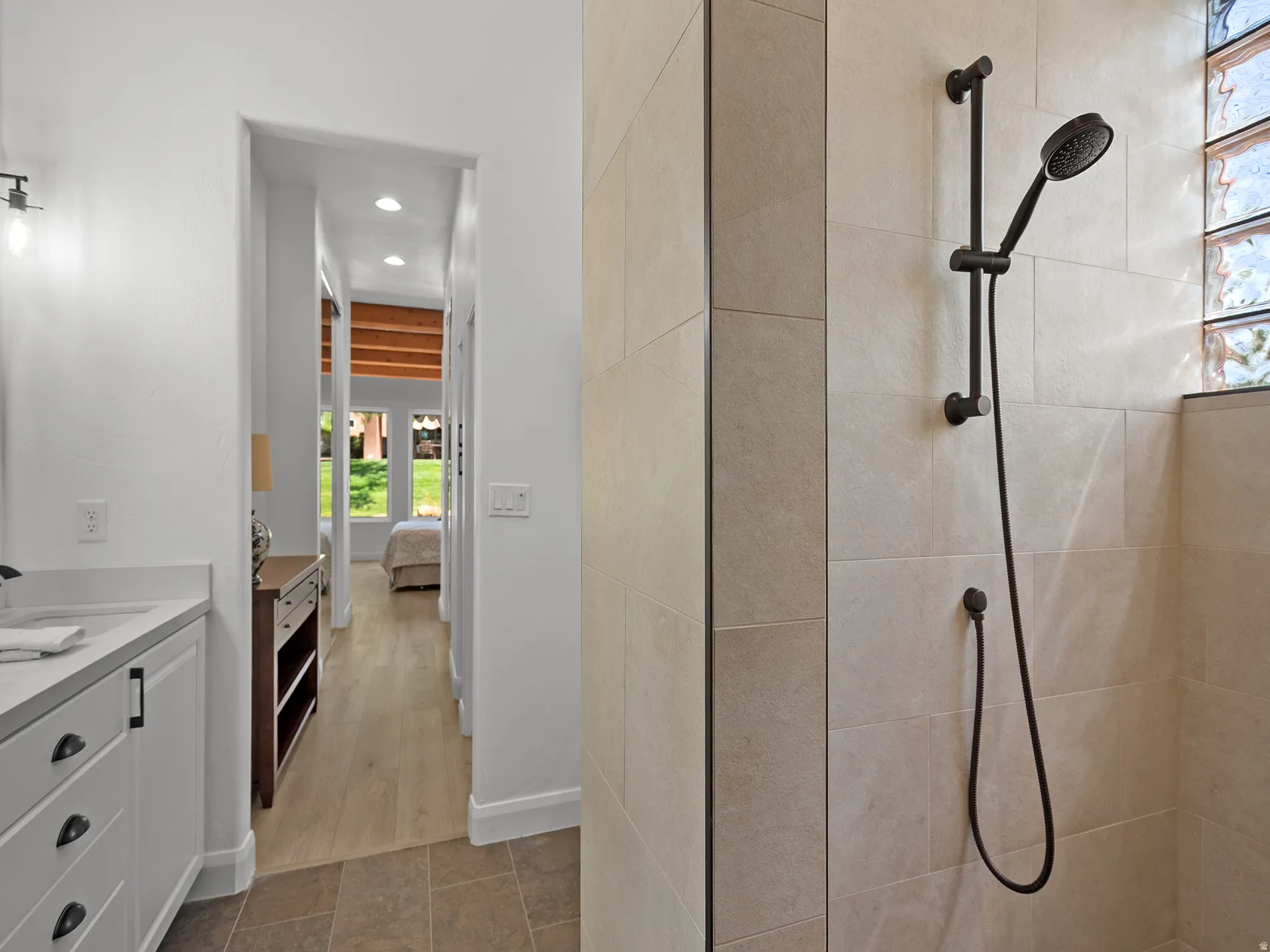 Walk-in shower with large format tile and black-finish fixtures, glass block window, and vanity with stone countertop