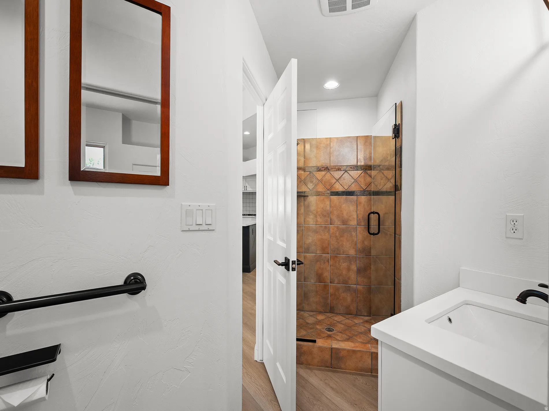 Walk-in shower with decorative tile, white vanity with integrated sink, matte black faucet, and wood-finish flooring