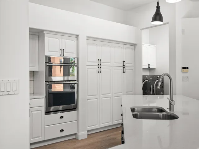 White cabinetry with black hardware, stainless steel double wall oven, integrated kitchen island sink with gooseneck faucet, white solid surface countertops, and wood-finish flooring