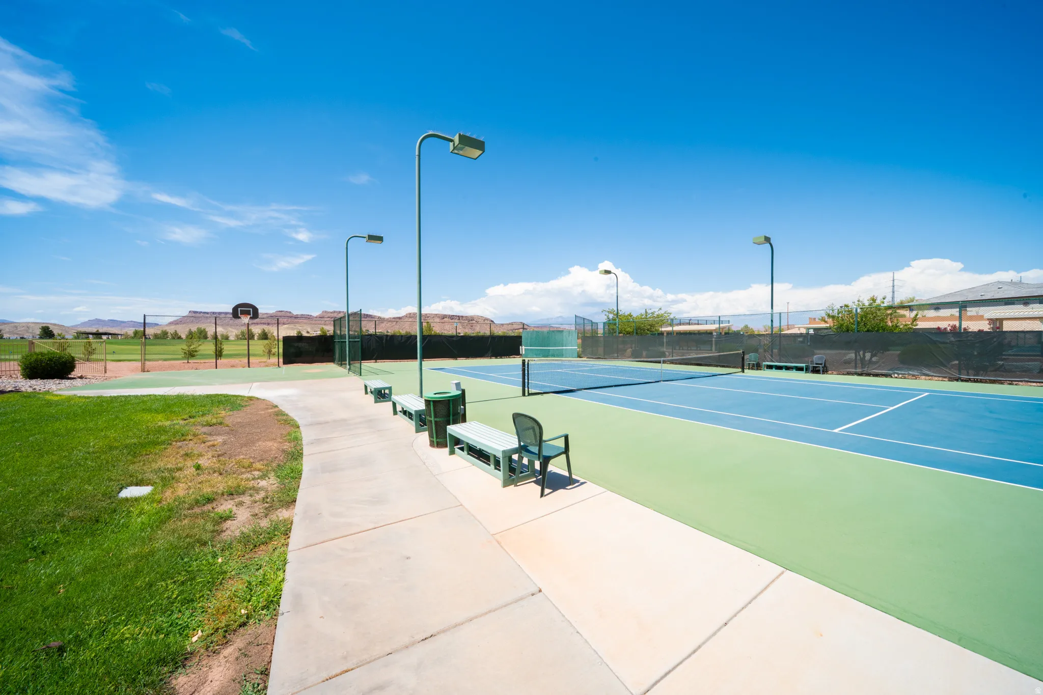 Paved walkway leading to two tennis courts with surrounding fencing, overhead lighting, and benches