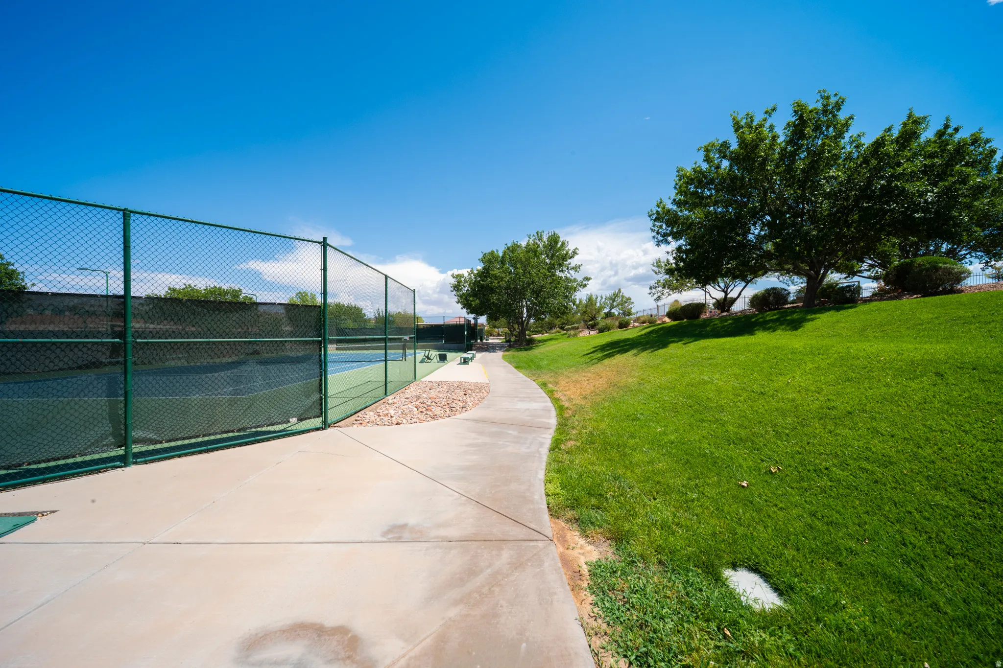 Paved pathway alongside a fenced tennis court, complemented by a manicured grassy slope and mature trees