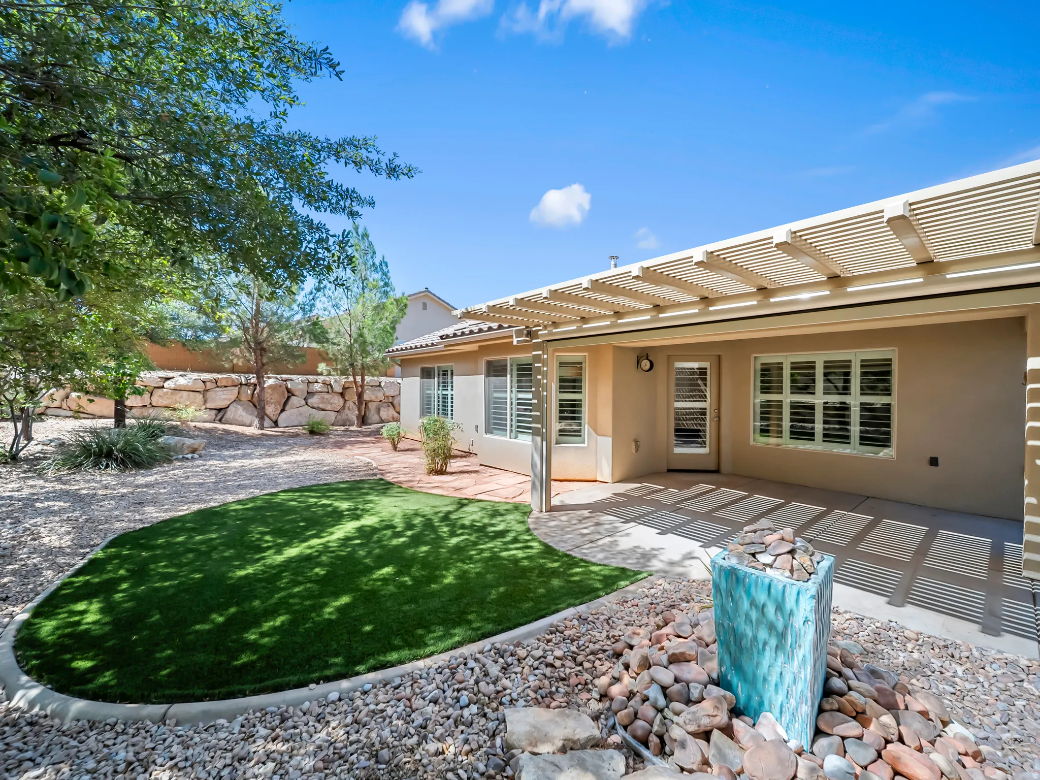 Rear patio with a slatted pergola, artificial turf lawn area, rock landscaping, and a decorative water feature