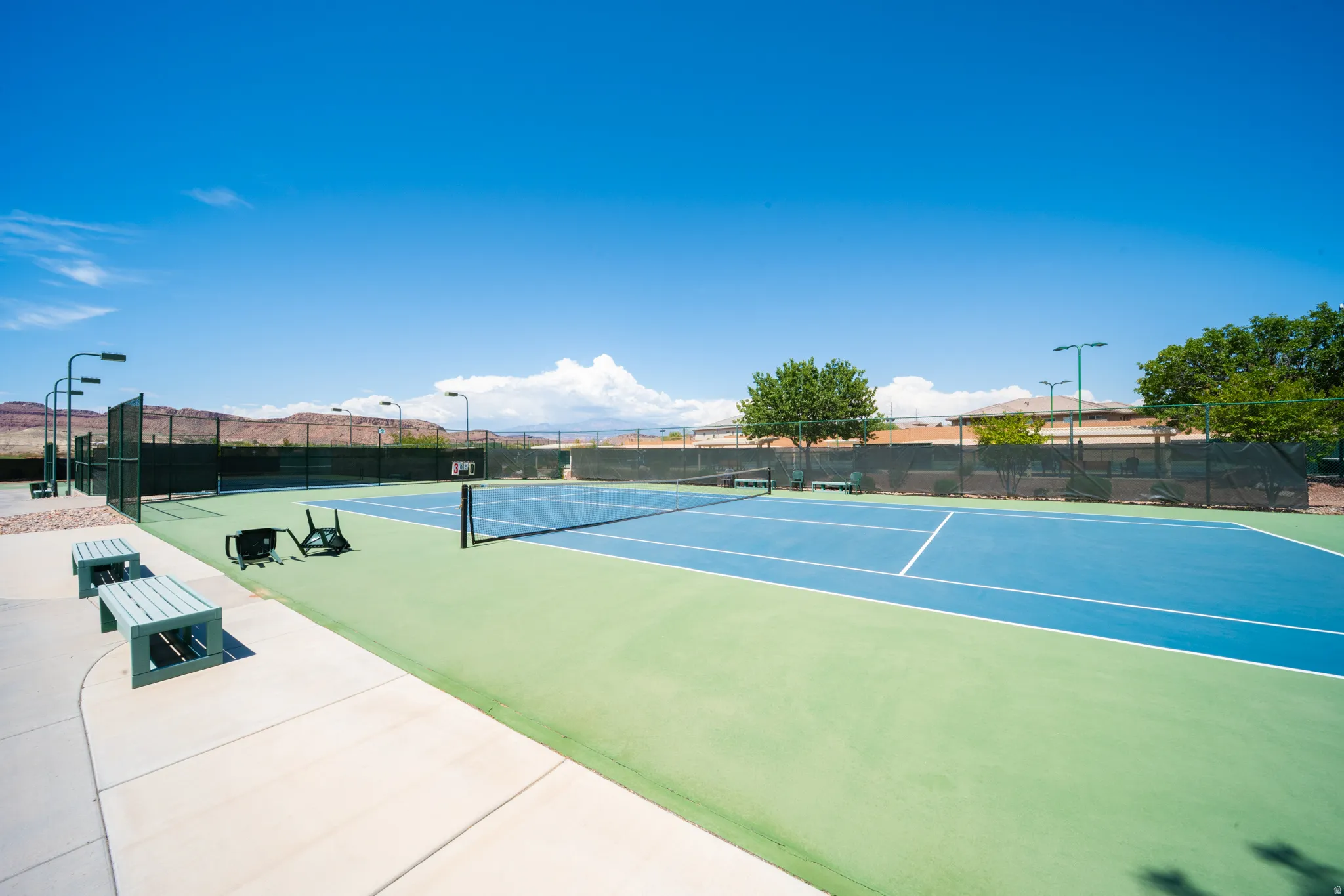 Full-size tennis court featuring a blue playing surface with green surrounds, perimeter fencing, and overhead lighting