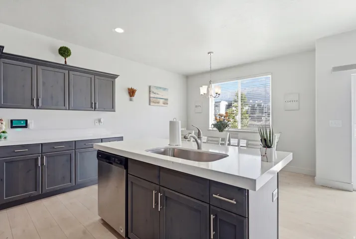Kitchen island with an undermount sink and stainless steel faucet, dark wood-finish cabinetry, light-toned countertops, stainless steel dishwasher, and light wood-finish flooring