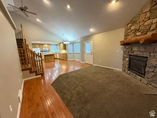 Vaulted ceiling with a ceiling fan, stone-clad fireplace with a wood mantle, wood-finish flooring, and natural wood staircase