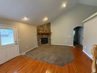 Vaulted ceiling, stone fireplace with log mantle, recessed lighting, wood-finish flooring, and arched doorway