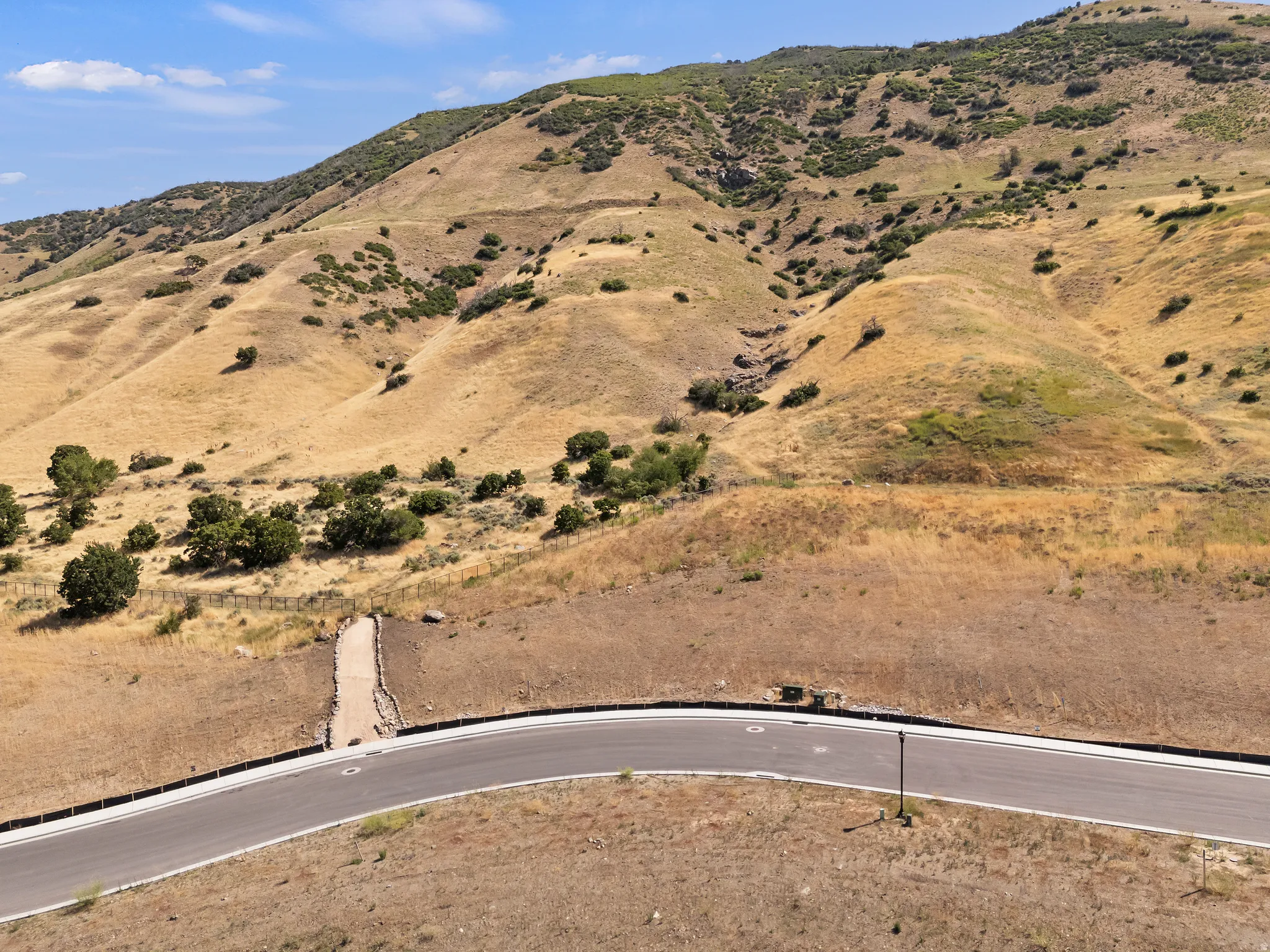 Paved road with marked lanes and curbs, bordering an expansive hillside with varied terrain