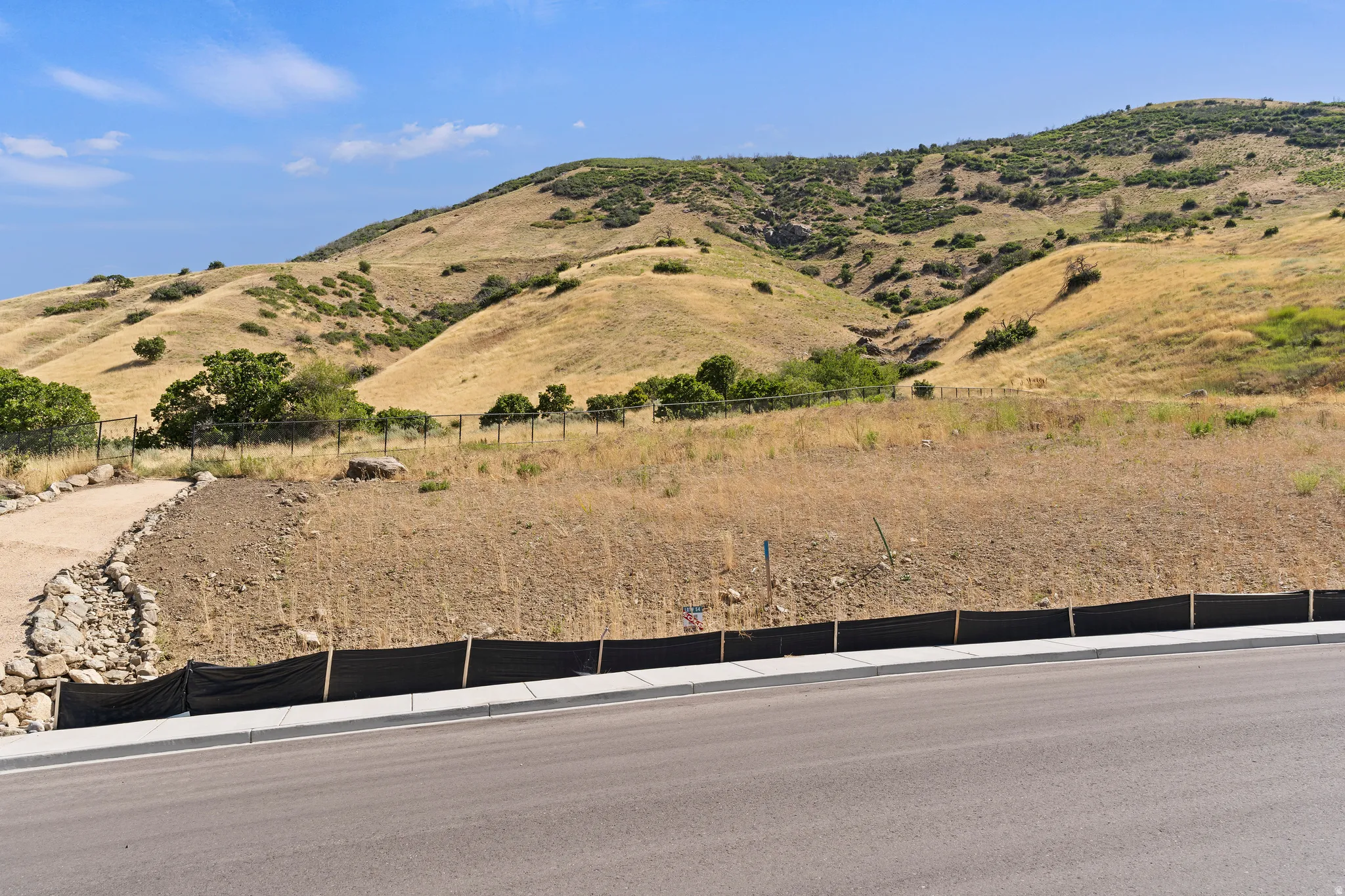 Paved road frontage with sidewalk and curb, featuring a graded lot with a partial rock retaining wall and natural hillside backdrop