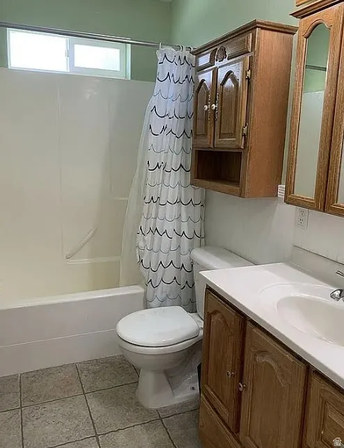 White bathtub with integrated shower, white toilet, tile flooring, and wood-finish vanity with an integrated sink