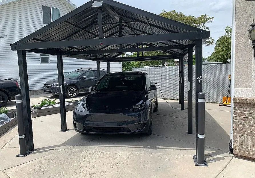 Paved driveway with a metal carport featuring a corrugated roof, integrated lighting, and multiple support columns