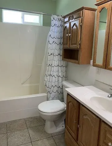 White bathtub with integrated shower, white toilet, tile flooring, and wood-finish vanity with an integrated sink