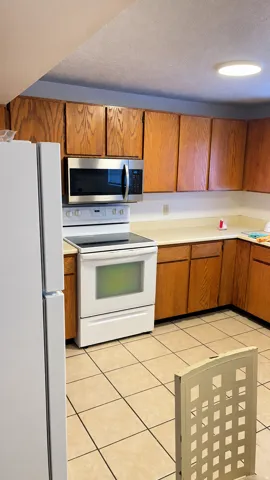 Kitchen featuring wood cabinetry, light-toned countertops, tile flooring, a stainless steel microwave, and white appliances