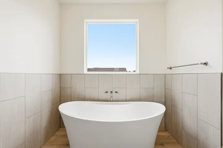 Freestanding soaking tub with wall-mounted fixtures, wood-finish flooring, and light-toned wall tile