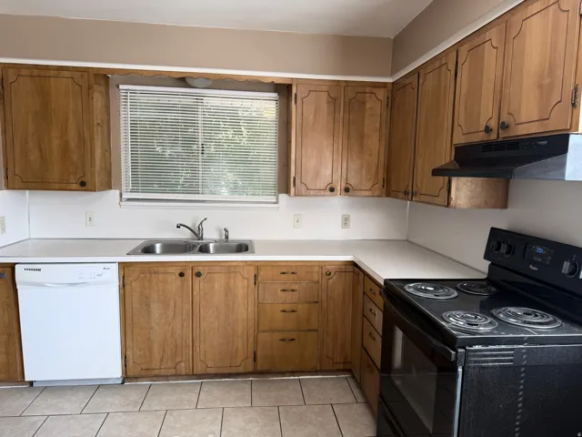 Kitchen featuring wood-finish cabinetry, light-toned countertops, a double basin stainless steel sink, a white built-in dishwasher, and a black electric range with an overhead exhaust fan