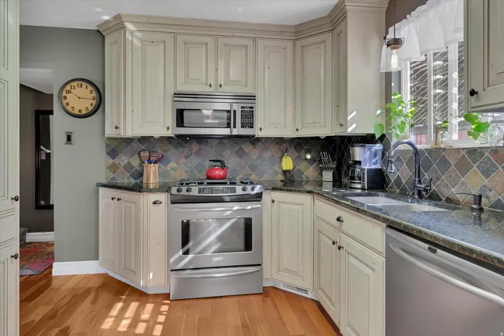 Hardwood flooring, distressed finish cabinetry, stainless steel appliances, dark stone countertops, and a diagonal tile backsplash