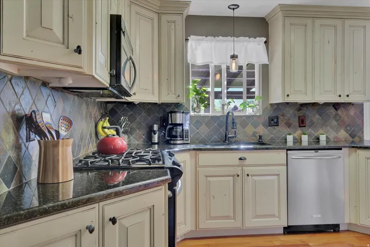 Cream-finish cabinetry with dark hardware, dark stone countertops, tile backsplash, stainless steel dishwasher, and a built-in microwave