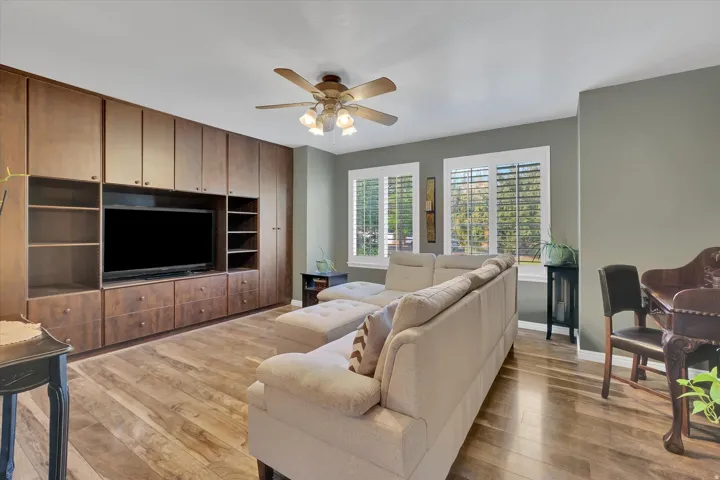 Built-in media center with extensive wood-finish cabinetry, wood-finish flooring, two windows with plantation shutters, and a ceiling fan with integrated lighting