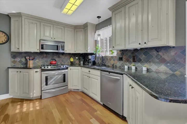 Kitchen with wood-finish flooring, light-toned cabinetry, stainless steel appliances, dark stone countertops, and a multi-color tiled backsplash