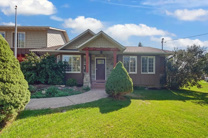 Stone accent columns, covered front entry, bay window with plantation shutters, mature landscaping, and a manicured lawn