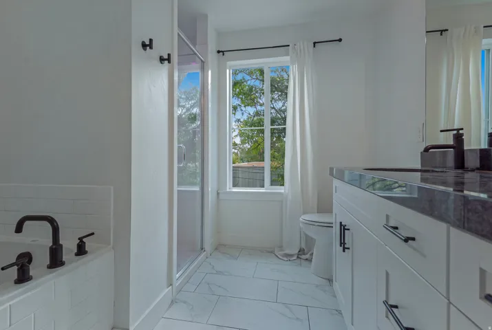 White tile flooring, built-in bathtub with subway tile surround, glass-enclosed shower, white vanity with stone countertop, and a window with natural light