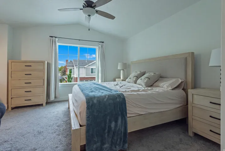 Vaulted ceiling with a ceiling fan, large window with white trim, and light gray wall-to-wall carpeting