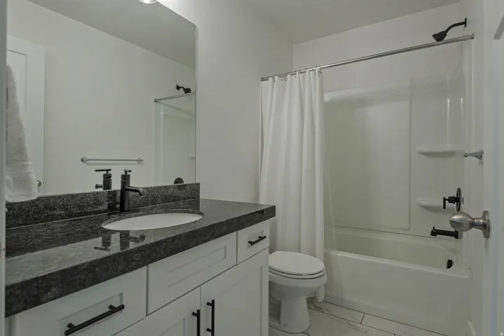Vanity with a dark countertop and undermount sink, matte black faucet, white cabinetry, and a white tub with shower