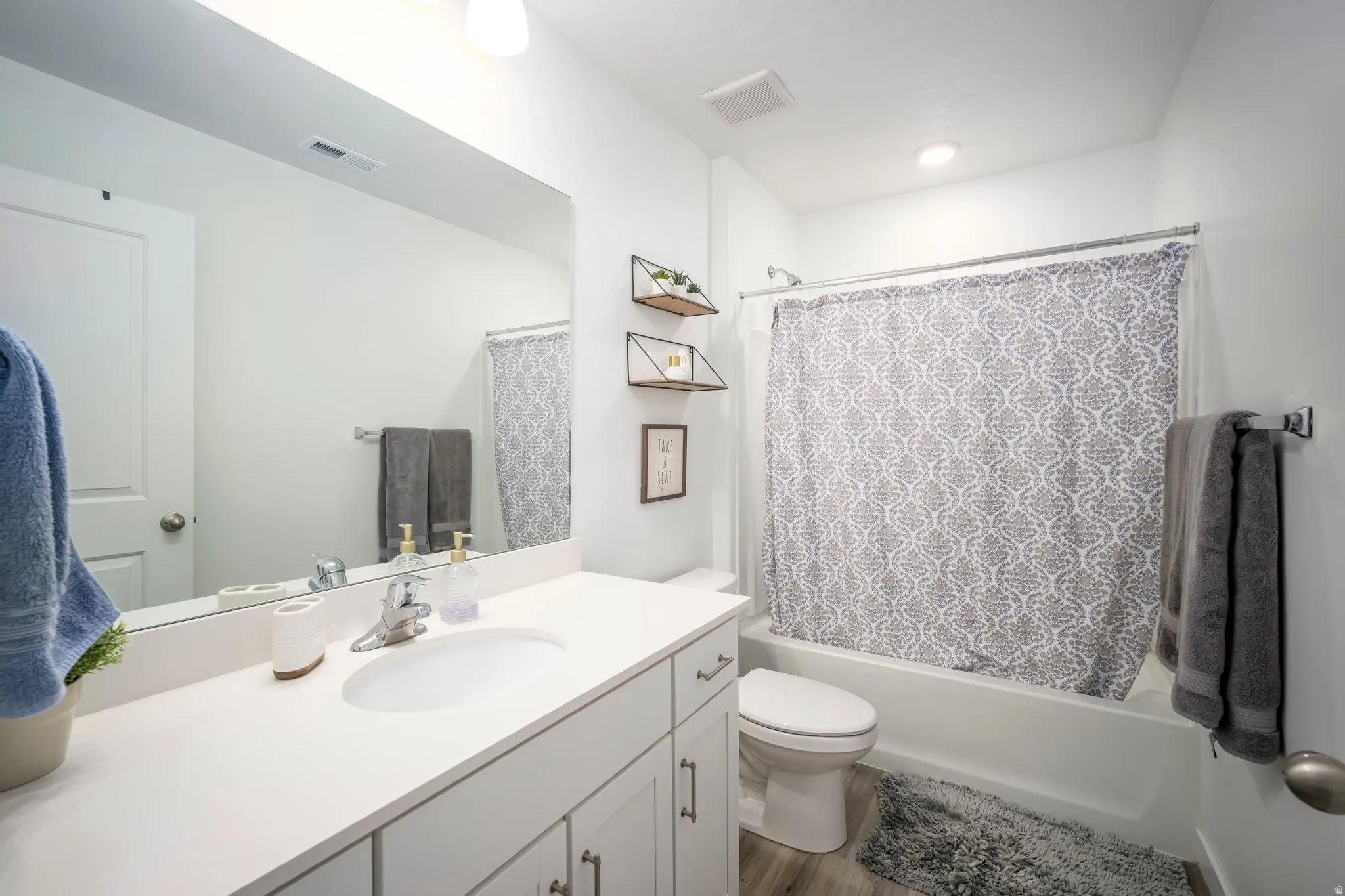 Vanity with white countertop and integrated sink, chrome faucet, white cabinetry with silver hardware, full-width wall mirror, and a white tub with shower