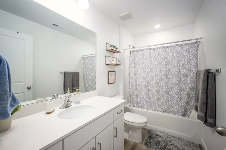 Vanity with white countertop and integrated sink, chrome faucet, white cabinetry with silver hardware, full-width wall mirror, and a white tub with shower