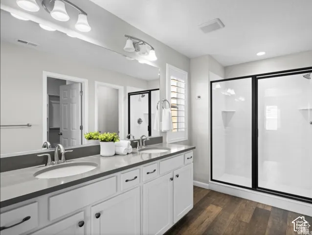 Dual vanity with undermount sinks, spacious shower with black-framed glass enclosure, wood-finish flooring, and white cabinetry with dark hardware