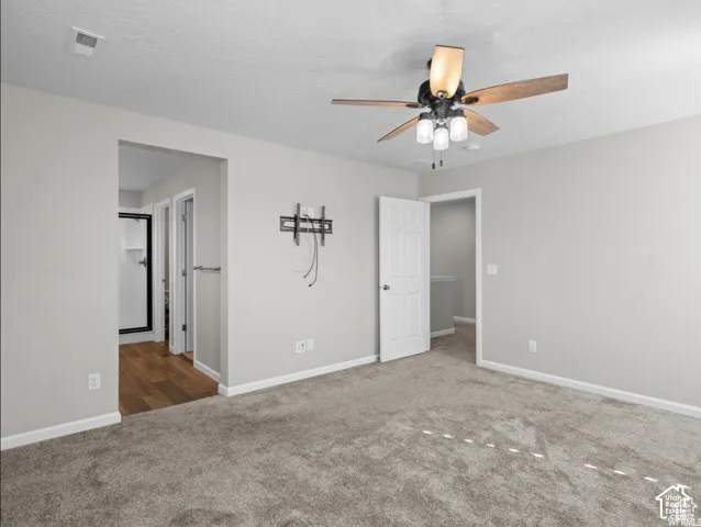 Carpeted flooring with light neutral wall paint, ceiling fan with integrated lighting, wood-finish flooring visible through doorway, and a white panel door