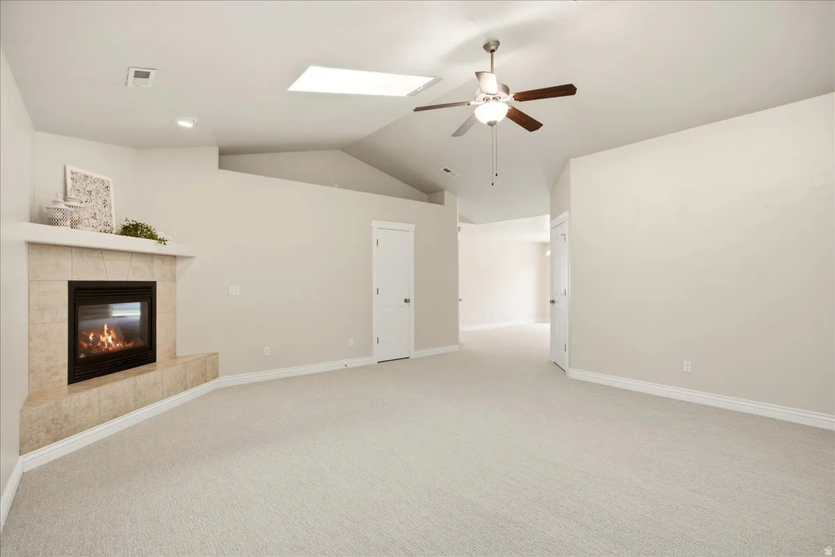 Carpeted room featuring a tiled fireplace, vaulted ceiling, ceiling fan with light fixture, and a skylight