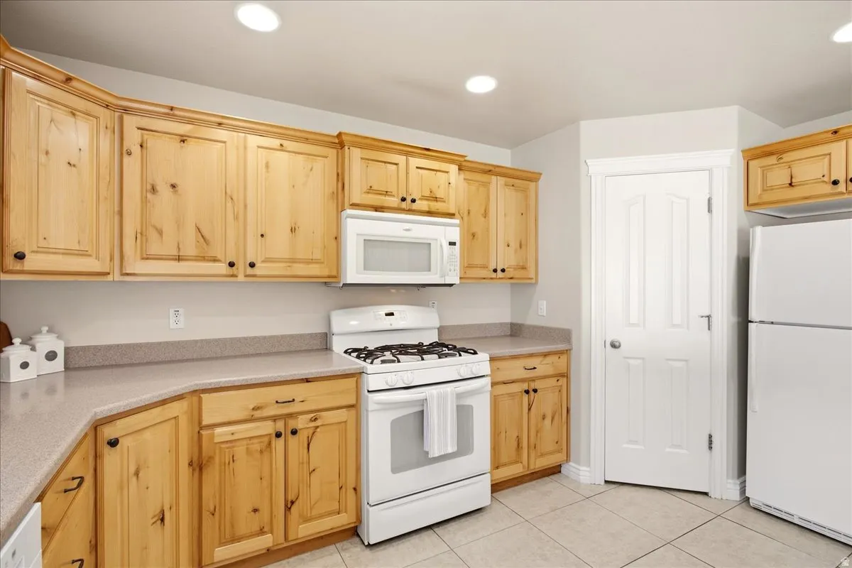 Wood-finish cabinetry with black hardware, white appliances, solid surface countertops, recessed lighting, and tile flooring