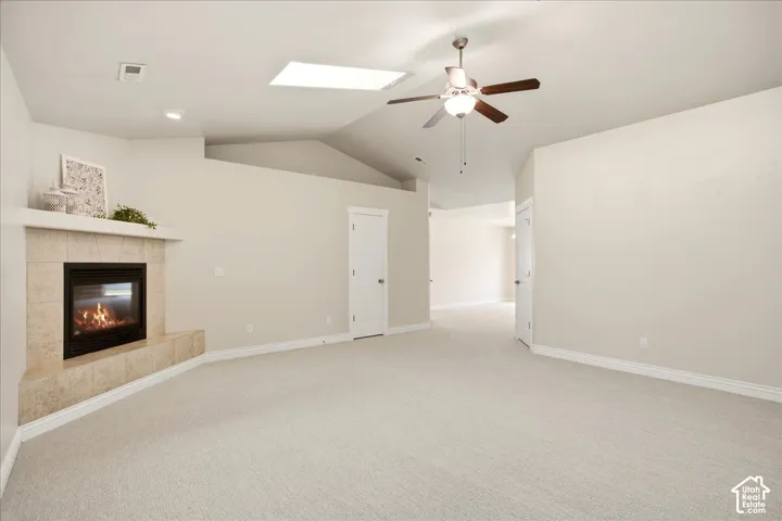 Carpeted room featuring a tiled fireplace, vaulted ceiling, ceiling fan with light fixture, and a skylight