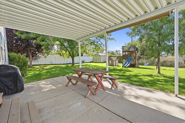 Covered patio with concrete flooring, supported by white posts, overlooking a fenced backyard with mature trees