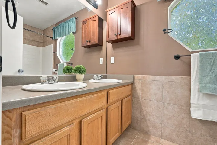 Dual sinks set in a stone-look countertop, wood cabinetry, and an octagonal window with decorative glass