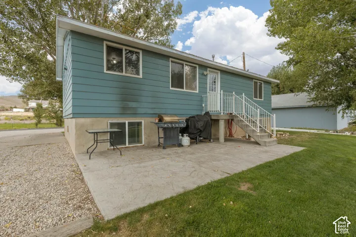 Exterior featuring blue siding, a concrete patio, a concrete staircase with white railings, a basement-level window, and a gravel driveway