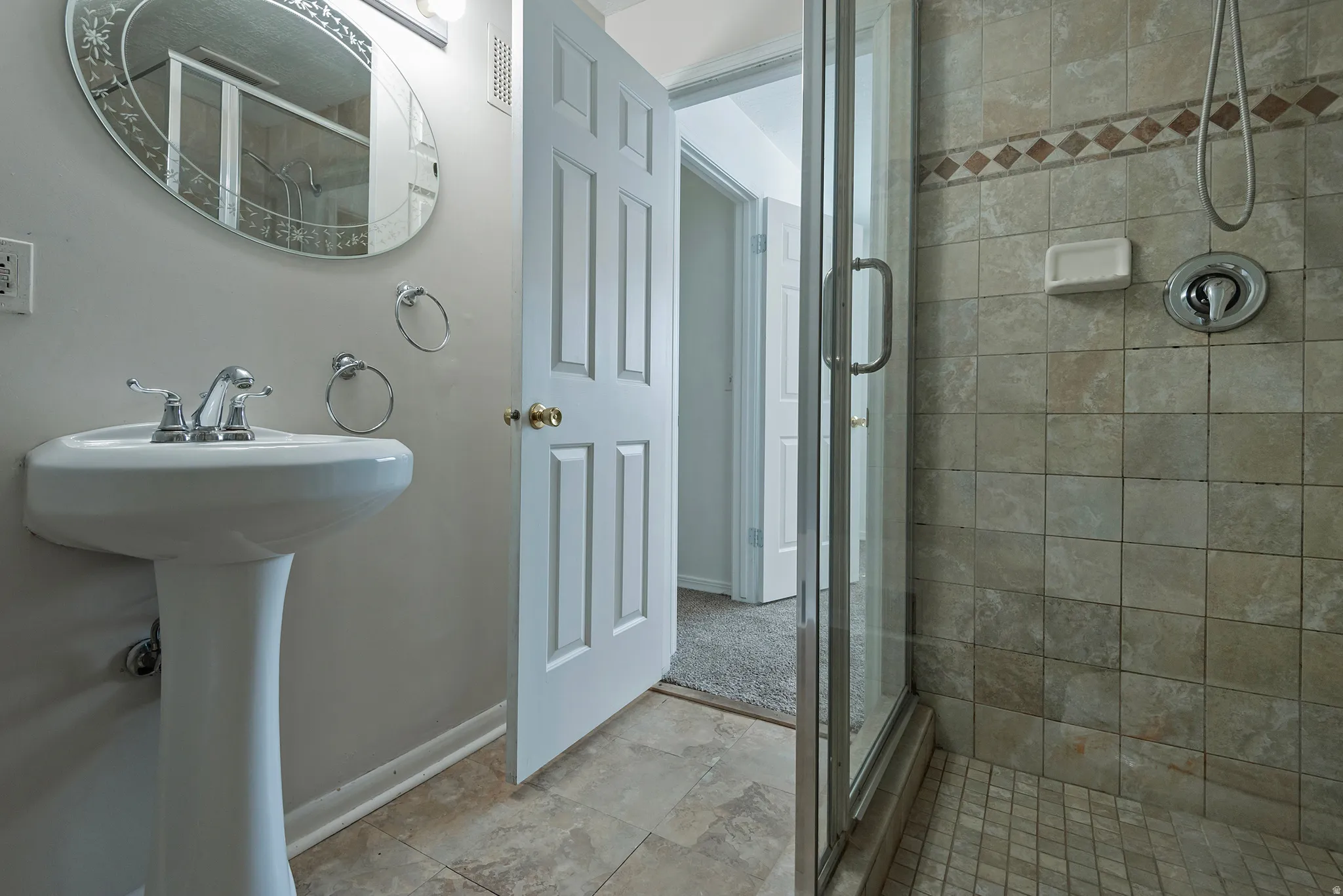Pedestal sink with chrome faucet, oval mirror, walk-in shower with glass enclosure, tiled shower surround, and tiled flooring